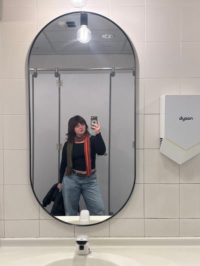 The image depicts a female individual taking a mirror selfie in a public restroom. The exact location within Sesto Fiorentino, Italy, is not identifiable from the visual evidence.
The central subject is a young adult female, reflected from the waist up in a large, vertically oriented oval mirror with a black frame. She has medium-length brown hair with bangs and is wearing a long-sleeved black top, light blue loose-fitting jeans, a dark belt, and a multi-colored striped scarf with red, orange, green, and brown hues draped around her neck. In her raised right hand, she holds a light-colored smartphone with a clear, MagSafe-compatible case, positioned to capture her reflection. Her left hand holds a folded dark-colored jacket or coat. Her gaze is directed towards the smartphone/mirror.
The background, visible both directly and in the mirror's reflection, consists of walls clad in large, rectangular white tiles with light grey grout. To the right of the mirror, a white Dyson brand hand dryer with a gold or tan base is mounted on the tiled wall. Below the mirror, a white ceramic sink basin is visible, equipped with a white, modern-style faucet featuring a black top. Reflected in the mirror are two light-grey or white public restroom stall doors with silver horizontal rails and a central hinge. The ceiling, also reflected, appears to be a light-colored grid ceiling with several embedded circular lights and a smoke detector. A bare incandescent light bulb in a black fixture is suspended from the ceiling, visible above the mirror's top edge. The lighting is bright and appears to be artificial overhead illumination.