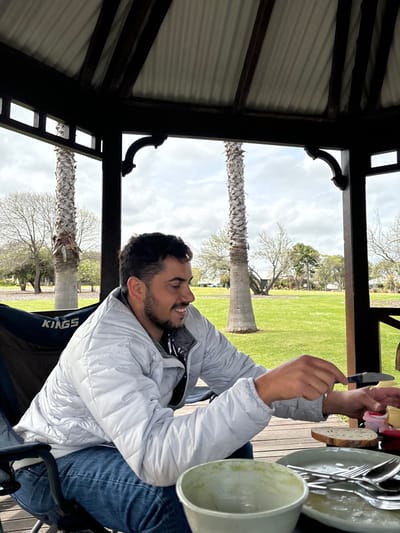 The image depicts an adult male seated within a dark-stained wooden gazebo in a public park in Margaret River, Australia. The gazebo features a corrugated metal roof and decorative curved timber supports. The male, appearing approximately 25-35 years old with dark hair and a short beard, wears a light grey quilted puffer jacket over a dark top, and blue jeans. He is seated in a black camping chair, partially visible, which bears a 'KINGS' brand logo. He holds a small knife in his right hand, actively engaged with food on a table in front of him. On the table, there are multiple ceramic plates with cutlery, slices of rustic bread, and small individual colorful containers of spreads or condiments (pink, yellow, and red). A light green ceramic bowl, appearing used, is positioned in the lower left foreground. The background displays a vast green lawn, several tall palm trees with distinct ringed trunks, and various deciduous trees, some bare, under an overcast sky. Faint structures are discernible in the far distance at the edge of the park.