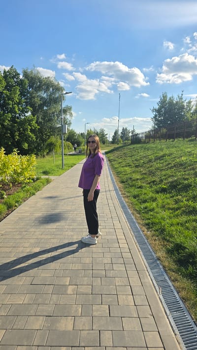 A woman stands on a paved walkway in a park in Razvilka, Russia. The woman is wearing a purple t-shirt, black pants, and white sneakers. She has sunglasses on and her hair is brown. She is facing the camera and appears to be smiling. The walkway is made of grey paving stones. To the left of the walkway is a grassy area with trees and bushes. To the right of the walkway is a grassy slope with a fence at the top. The sky is blue with some white clouds. There is a street lamp on the left side of the walkway and another one further in the distance. There are two other people walking in the distance in the park.