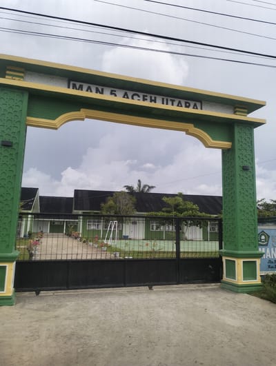The image shows the entrance gate to a school in Muliengmanyang, Indonesia. The gate is green and yellow, with an inscription on the top arch that reads "MAN 5 ACEH UTARA". Through the gate, several green school buildings with black roofs are visible, along with a black metal fence. The ground in front of the gate is paved with concrete. The sky is overcast. There are some potted plants and flowering bushes near the fence. A sign with the school's emblem is visible on the right side of the gate.