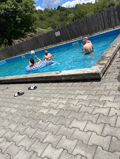The image depicts an outdoor swimming pool on a sunny day. In the pool, three individuals are present: two men and one woman. One man is lying on a colorful inflatable shaped like a mermaid's tail, while another man is standing and tossing a volleyball. The woman is standing next to the man who is tossing the ball. The pool is surrounded by a wooden fence and a concrete deck paved with interlocking stones. Lush green trees and vegetation are visible in the background, suggesting a natural or semi-rural setting. A sign with text is affixed to the wooden fence. There are two pairs of flip-flops on the paved area near the pool. The scene appears to be at the swimming pool facility in Liava Reka, Bulgaria.