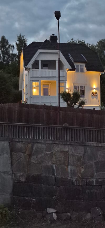 The image depicts a white, two-story house with a dark roof, illuminated by warm outdoor lights. A dark wooden fence runs along the front of the property, behind which a stone wall is visible. A street lamp stands in front of the house. A sign on the house reads "29 TL. SALCS EKSTRE." The scene appears to be set in a residential area at dusk. The house is nestled amongst trees, and the sky is cloudy.