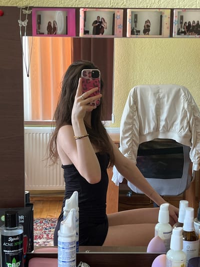 A young woman with long brown hair is taking a mirror selfie in what appears to be a bedroom. She is wearing a black strapless dress and has on a ring and bracelets. In the reflection, on the wall above her, are several framed photographs of people. The room has a wooden floor and a radiator is visible behind her. In the foreground, on a surface in front of the mirror, are various cosmetic products.