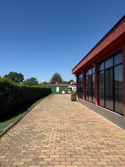 The image depicts a paved walkway leading past a red-colored building with large dark-tinted windows. To the left of the walkway, a meticulously maintained green hedge runs along the edge of the property. In the background, several trees and smaller green structures are visible under a clear blue sky. The walkway is constructed of interlocking pavers in shades of red and yellow. The scene appears to be an outdoor area associated with a building, possibly a commercial establishment or event venue, given the formal landscaping and building design. The location is identified as Yagoda, Bulgaria. There are no people or discernible actions taking place in the image.