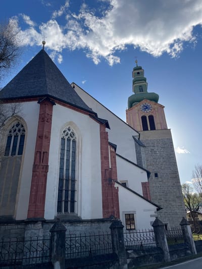 The image features the Chiesa di Santa Maria Assunta (Church of Saint Mary of the Assumption) in Sterzing - Vipiteno, Italy, captured from a low angle. The church exhibits a combination of architectural styles, with a white facade accented by red brick details. The roof is dark gray. Gothic-style windows are visible. The church tower, partially in view, displays a clock face and a green onion dome atop a pink and gray structure. The sky is blue with scattered white clouds. A wrought iron fence and the bare branches of trees are present in the foreground.