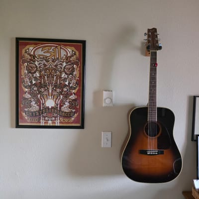 An acoustic guitar is hanging on a wall in a room. The guitar is sunburst brown and black, with a Fender headstock. A guitar pick is lodged between the strings on the headstock. To the left of the guitar, a framed poster is hanging on the wall. The poster is red and gold and appears to be a concert poster. Below the poster is a white thermostat. Below the thermostat is a white light switch. The wall is textured and painted a light color. The scene is photographed from a medium distance.