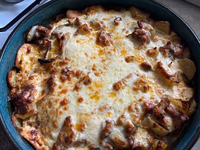 A high-angle, close-up photograph displays a freshly baked casserole dish in a round, dark teal or deep blue pan. The dish contains layered ingredients, predominantly wedge-cut potatoes, visible along the perimeter and interspersed throughout, some retaining their skin and showing golden-brown edges. Chunks of browned meat, possibly chicken or pork, are scattered among the potatoes. Distinct mushroom pieces, including one with visible gills on its underside, are identifiable. The entire surface is covered by a melted cheese layer, characterized by irregular browning and bubbling, with glistening rendered fat visible in several areas. The pan rests on a light-colored surface, likely a kitchen countertop or stovetop, with a partial view of a white ceramic or enamel surface and a black burner grate element in the upper left background. The scene is located in Gorna Rositsa, Bulgaria. No individuals or active interactions are depicted.