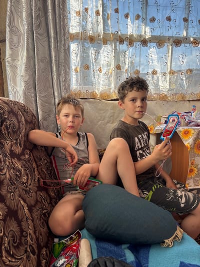 This image captures two young boys indoors, likely in a home in Маркидоновка, Russia. The scene appears to be during the daytime, with sunlight streaming through a patterned sheer curtain on the left. The boys are seated on a patterned sofa.

The boy in the foreground is lying on his side, looking towards the camera. He is wearing a grey tank top with black and red trim and a number "1996" printed on it. He holds a green and pink toy with a tube, possibly for blowing bubbles, in his right hand, with the tube near his mouth.

The older boy is sitting behind and to the right of the foreground boy. He is wearing a dark t-shirt with white lettering and patterned shorts. He is also holding a similar toy with a red tube, with the tube near his mouth, and looking directly at the camera. The toys appear to be bubble wands with connected pouches.

In the background, a wooden table is visible with a floral tablecloth and some items on it, including what looks like packaged drinks or snacks. The overall atmosphere seems relaxed and casual, suggesting a moment of playtime or leisure. The sunlight creates a warm glow in the room, and the boys' expressions are neutral.
