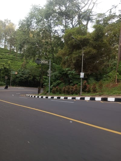 The image depicts a road in Taunggyi, Myanmar. The road is asphalt with a yellow line marking the center. A curb is painted with alternating black and white blocks. Lush green vegetation covers the hillside. A street lamp is visible on the right side of the road. A motorcycle is at the intersection in the background. Road sign is visible over the road.