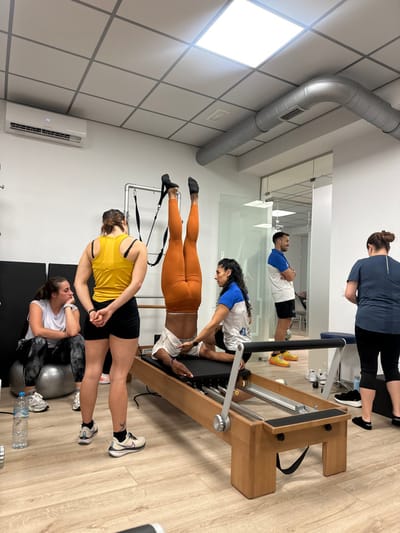 A group of people are in a Pilates studio, some working on a Pilates reformer machine and other equipment. One person in orange leggings lies on the machine while a trainer adjusts the straps. Another person in a white shirt with a ball behind them does a different exercise. There is a man in white and blue lying on the machine as well. The Pilates reformer has wooden panels and the room has white walls.