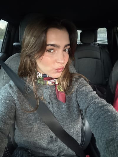 A woman with brown hair and light skin sits in a car, wearing a gray cardigan. She is secured with a seatbelt. A colorful scarf adorns her neck. The interior of the car features black seats and panels. The image is taken from a slightly angled perspective, close to the woman's face, and appears to be a selfie.