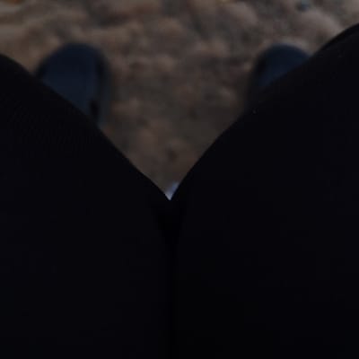 The image displays a first-person, downward-facing perspective of an individual's lower body in Sámara, Costa Rica. The subject's legs, clad in dark, possibly black, ribbed fabric, occupy the foreground and midground, extending from the bottom of the frame and converging towards the center before diverging. A blurred view of two dark shoes or feet is visible at the distant end of the legs. The ground beneath and between the legs appears to be an irregular, textured surface of brown earth or sand, with scattered darker particles, suggesting a natural outdoor setting such as a path or beach. The lighting is subdued, contributing to the dark tones of the clothing and slightly dimming the ground. No other individuals or specific objects are identifiable. The scene depicts a stationary posture, likely standing or sitting, looking down at the feet and immediate surroundings.