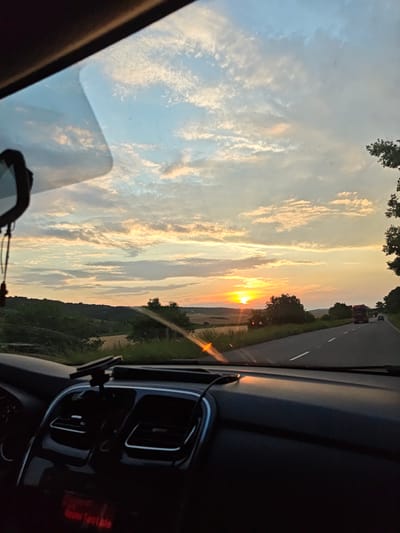 The image captures a view from inside a car driving on a highway towards a sunset. The sky is filled with scattered clouds tinged with orange and yellow hues from the setting sun, which is visible on the horizon. Rolling hills covered in green foliage and fields are seen on the left side of the road, with trees lining the right side. A red truck and other vehicles are driving ahead on the asphalt road. The car's dashboard, including air vents and a phone mount, is visible in the foreground. The scene is set in Zakharivka, Ukraine.