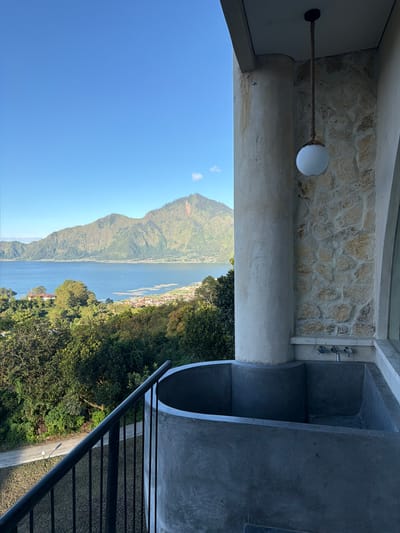 From an elevated, partial indoor vantage point, a view opens to the landscape of Kedisan, Indonesia. The sky is clear and blue. A mountain stands prominently in the background. A body of water stretches out below the mountain. Along the shores, a settlement of buildings are visible, nestled among trees. Lush green trees and vegetation occupy the land below. The perspective is from an outdoor space, where construction materials like stone are used. A white spherical light fixture hangs from the ceiling. To the right, a dark gray element resembling a bath is constructed. Stairs with black metal railings are seen. The entire scene gives an impression of a view from an elevated location, possibly from a unique architectural viewpoint overlooking a natural landscape.