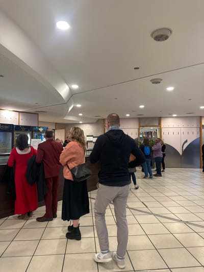 The image shows a group of people standing in line in a large, open room with a tiled floor and a high ceiling. There are several ceiling lights, a white ceiling, and a series of large, double doors at the far end of the room. There are several people standing in line in the foreground of the image, and another group of people standing further down the hallway. The people in the line are all facing the same direction, presumably towards the doors at the end of the hallway. The group at the back of the hallway is talking to each other. The people in line are all wearing casual clothing. There is a large rectangular counter in the background with refrigerators behind it. The image was taken in Bingen am Rhein, Germany (latitude: 49.969, longitude: 7.892). The location is likely a restaurant or a cafe.
