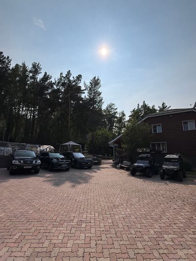 The image depicts an outdoor scene in Sukhaya, Russia, on a sunny day with a clear blue sky. In the foreground, a red brick paver driveway is visible. Parked on the driveway are several vehicles, including a black Nissan Patrol SUV on the left, followed by a black Mercedes-Benz SUV, and another black SUV on the right. Further to the right, two off-road vehicles, possibly ATVs or UTVs, are parked. In the background, a wooden house with a dark brown exterior and white-trimmed windows stands. Tall pine trees line the edge of the property, casting shadows on the driveway. A white gazebo is also present near the center of the frame. The overall impression is that of a private residence or a recreational property. There are no people visible in the image.