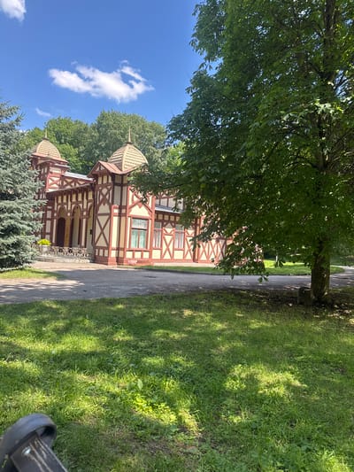 A light beige and red half-timbered building with peaked roofs is situated in a park on a sunny day. The building has decorative trim and arched entrances. In the foreground, a large green tree with sparse leaves casts dappled shadows on the grass and a paved path. To the left of the building, a tall, blue-green conifer tree is visible. The sky is bright blue with scattered white clouds. The image appears to be taken in Yessentuki, Russia, likely within a park or sanatorium. No people or animals are visible in the frame.
