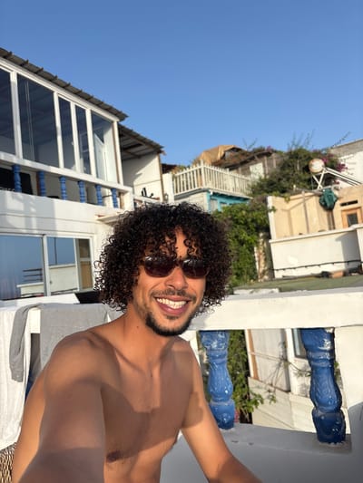 A man with curly dark hair and sunglasses smiles at the camera while sitting on a balcony. He is shirtless and appears to be in a tropical or coastal environment, with buildings and lush greenery in the background. The scene suggests a relaxed vacation or a casual outdoor setting.