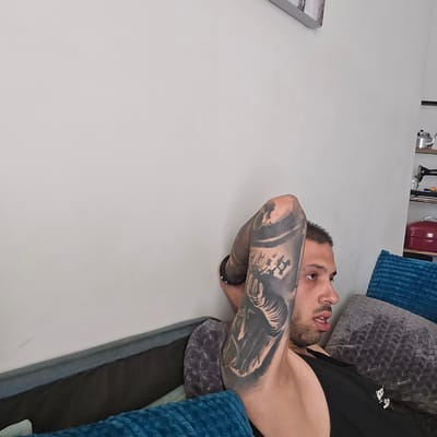 The image shows a close-up of a man with a large tattoo on his right arm, lying on a sofa. He is wearing a black tank top and has his arm raised behind his head. The man appears to be in his late twenties or early thirties, with short dark hair and a stubbled chin. He has his eyes open and is looking to his right, with his mouth slightly agape. The sofa is dark gray and is covered with several pillows, one of which is bright blue and textured. In the background, a white wall is visible, and to the right, a shelf with a teapot, a sewing machine, and a red tin can can be seen. The lighting is soft and natural, suggesting an indoor setting. The image does not provide any specific information about the location in the city of Agii Anargiri, Greece.