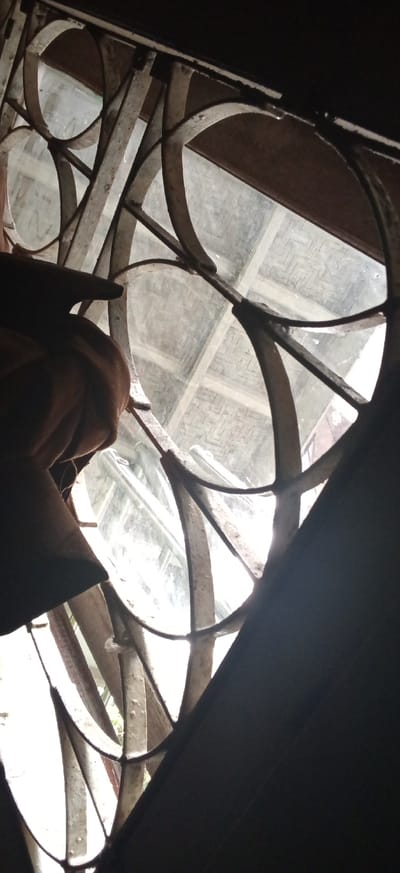 The image captures an interior view looking outwards through a window, characterized by a dark, weathered wrought iron grill. The grill features an ornate, geometric design composed of intersecting curved and straight metal bars, creating circular and abstract openings. Some of the glass panes behind the grill appear dirty or frosted, while others offer a clearer, though still hazy, view of the exterior.
Through the window, a brightly lit outdoor scene is visible, backlighting the entire structure and suggesting daytime. A distinctive textured surface, possibly a woven bamboo or rattan material, or a patterned roof or wall, is discernible through the clearer sections of the glass. In the foreground, primarily on the left side, a dark brown, somewhat rumpled fabric object is visible, held by what appears to be a human hand or fingers. This fabric partially obscures the lower-left section of the window and the grill. The overall scene is high-contrast, with the interior elements and the metalwork appearing in silhouette against the bright light from outside. No discernible text is visible within the image. The setting is in Banjaran, Indonesia.