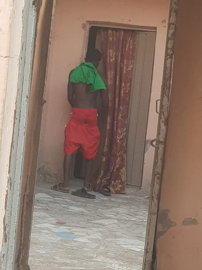 A person wearing red shorts and a green head covering is inside a doorway of a building with light peach walls. The doorway has a metal roll-up door partially lowered, behind which a patterned curtain is visible. The person is wearing sandals. The floor is composed of patterned tiles. The location is in a city, potentially Dutsinma A, Nigeria.