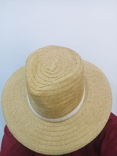 A light-colored, wide-brimmed straw hat is depicted, positioned as if worn by an individual. The hat is constructed from woven straw in a light beige or tan hue. Its flat-topped crown features a prominent spiral or concentric circular weave pattern at its center. A white ribbon band encircles the base of the crown, separating it from the wide, textured brim. A portion of a dark red or maroon garment is visible beneath the hat's brim, indicating it is likely worn by a person. The background consists of a plain, light grey or off-white surface, suggesting an indoor environment or a studio setting. The lighting is even and diffused. The scene is situated in Festac Town, Nigeria. No other individuals, subjects, objects, or explicit actions or interactions are discernible.