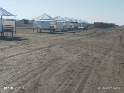 In Abilyatag, Azerbaijan, the scene depicts a sandy, outdoor area with a row of numbered white gazebos lining the beach. The gazebos are topped with white conical roofs and feature white paneling on the sides, with benches and tables inside. Some of the gazebos are marked with numbers, such as "6", "31", and "17". In the background, a dark, square-shaped structure is visible, along with a long train of what appear to be hopper cars. The ground is covered in sand with visible tire tracks. The sky is clear and blue. A timestamp in the lower right corner reads "03.07.2026 16:55".