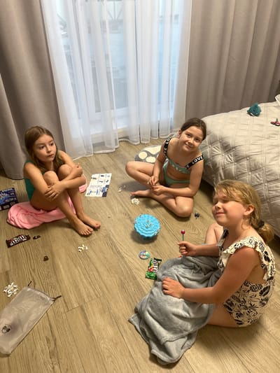 Three young girls are on the floor of a room in Новая Чернушка, Russia. The room contains a bed with a grey patterned comforter, curtains, and a wood-look floor. The girl closest to the camera is seated on a pink towel, hugging her knees. To her right is a box, candy, and a transparent bag of white toy pieces. Above her and to the right is another girl seated on a spotted pillow, wearing a two-piece swimsuit with black lettering. Next to her is a blue toy. The third girl is wearing a printed one-piece swimsuit and is partially covered with a grey blanket. Candy wrappers and small white toy pieces are scattered around the floor. The girls appear to be playing.