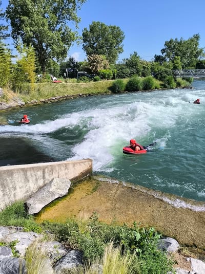 The image shows three people bodyboarding on the artificial whitewater course at Saint-Pierre-de-Bœuf, France. The location is identifiable by the prominent concrete structures and landscaping along the artificial course and the bridge in the background.

In the foreground, a person wearing a red helmet, a black vest, and fins is riding a red bodyboard in turbulent water near a concrete structure. Another person is further up the channel, while a third is visible in the distance. The water is a light green-blue color, with areas of white from the rapids.

The shoreline is lined with green vegetation, trees, and grass. In the background, there are more trees, a glimpse of a building, and a metal bridge spanning the water. The sky is clear and blue.