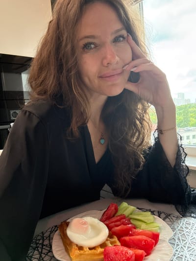 In this portrait, a woman with long, wavy brown hair and green eyes is captured from the chest up, leaning her head towards her hand. She is wearing a black robe with lace trim around the neckline. A delicate rose gold bracelet adorns her wrist, and a pendant necklace with a small light blue heart charm is visible at her décolletage.
To the right of the woman, a white plate holds a breakfast consisting of a waffle topped with a poached egg, sliced tomatoes, and sliced cucumber. The background suggests an indoor setting with natural light coming from a window, hinting at daytime. The exterior view through the window shows buildings and greenery, suggesting an urban or suburban environment. The overall mood appears relaxed and possibly serene, indicated by the woman's gentle pose and the intimate nature of the meal. The image was taken in Chashnikovo, Russia. No visible text is present in the media file.