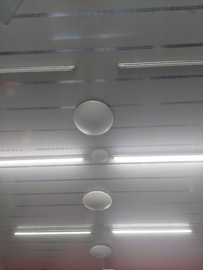 The image shows a close-up, overhead view of a modern, likely commercial or institutional ceiling. The ceiling is constructed of gray, horizontal panels, with thin, reflective metallic strips running horizontally between the panels, creating a striped effect.
The ceiling features several lighting fixtures. At the top of the image, two long, thin fluorescent tube lights are visible, running parallel to each other. Below these, a large, round, white ceiling fixture is centered. To the right of this fixture, a smaller, circular device, possibly a smoke detector or speaker, is attached to the ceiling.
Further down, two more long, bright fluorescent tube lights are installed horizontally. Between these, and slightly to the left, is another large, round, white ceiling fixture. Towards the bottom of the image, a portion of a fourth, similar round fixture is visible. The lighting from the fluorescent tubes creates bright, even illumination across the visible portion of the ceiling.
The image is taken from a low angle looking upwards, with no people or clear signs of the environment beyond the ceiling itself. The overall impression is one of clean, functional design, typical of modern indoor spaces. The location is identified as the City of Pematang Siantar, Indonesia.