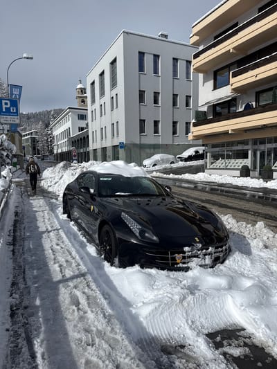 The image captures a snow-covered street scene in Sankt Moritz, Switzerland. A black Ferrari FF, identifiable by its badge and body style, is parked on the right side of the road, partially obscured by large piles of snow that extend onto its hood and roof. Icicles hang from the car's front grille.

To the left, a pedestrian walks away from the camera on a snow-lined sidewalk, wearing a light brown jacket with "GA" lettering on the back and dark trousers. Footprints are visible in the snow on the sidewalk.

The road surface in the center is wet and clear of deep snow, indicating recent clearing, but significant snow mounds line both sides, pushed against the curb. Further down the street on the right, two additional vehicles are visible, completely covered in snow.

Architecturally, the street is flanked by multi-story buildings. On the immediate right, a building with light beige balconies and dark railings features an "Engel & Völkers" real estate office at ground level. Across the street, a modern white building with numerous vertical rectangular windows is prominent. Further down, another white building with a dark, arched ground floor is visible. All buildings have snow accumulations on their roofs and ledges.

In the background, a distinct church tower with a domed roof stands out against a partly cloudy, light blue sky. Snow-covered mountains and evergreen trees are visible behind the urban structures. A streetlamp on the left features icicles. Street signs visible include a blue parking sign indicating "Zentrale Parkuhr" (Central Parking Meter) with hours "08:00 - 20:00 h" and a "max. 4 Std." (max. 4 hours) limit, as well as other regulatory signs further down the road.