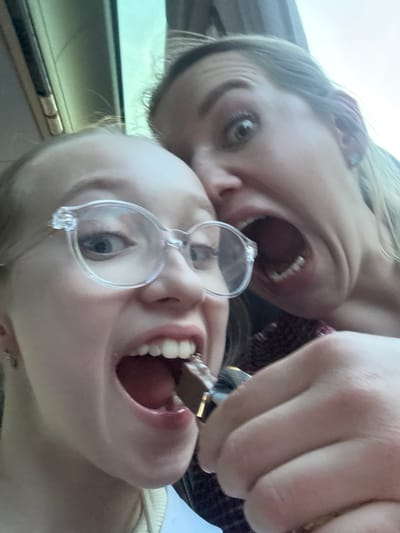 Two Caucasian women are in close proximity, possibly inside a vehicle. The woman in the foreground is wearing glasses and is in the process of eating a rectangular piece of chocolate. Her mouth is open. The second woman is directly behind her, also with her mouth open. The specific location in Lucca, Italy, is not discernible from the provided image.