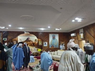 The image shows a room with a white ceiling and wooden walls. There are men in white and blue robes gathered in the room, some of whom are shaking hands. The men are gathered around a white couch and coffee table, all positioned near a window and a door. The room is decorated with paintings of people wearing traditional clothes, and ornate lamps hang from the ceiling. There are also numerous ornate, gold-colored wall decor pieces. It is a large room, with many features, including a large window on the right side of the frame. The coordinates provided, which are (latitude: 9.816, longitude: 11.314), place the image in Kaltungo, Nigeria.