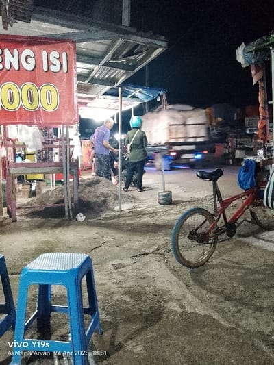 The image depicts a street scene in Kecamatan Kroya, Indonesia, at night, taken from a slightly elevated angle. The location is confirmed by the coordinates provided: latitude -6.422, longitude 107.996.
Two individuals are present. A man, wearing a light blue shirt and dark pants, stands near a stall. A woman, wearing a green jacket and a blue helmet, stands next to a motorcycle. Both individuals appear to be interacting with each other.
A red bicycle is parked in the foreground. The street surface is rough and appears to be made of concrete or asphalt. A pile of sand lies in the background, indicating construction or recent roadwork. A blue plastic stool sits in the foreground, suggesting an area of pedestrian traffic.
The image captures the hustle and bustle of everyday life in Kecamatan Kroya, highlighting the contrast between the bustling street scene and the stillness of the parked bicycle. The stall, the pile of sand, and the bicycle hint at local commerce and transportation. The time is likely evening or early night due to the dimly lit environment and the presence of streetlights.