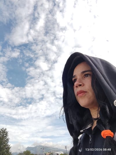 The image displays a female individual outdoors in Puembo, Ecuador, on 13/02/2026 at 08:48. The individual is positioned on the right side of the frame, observed from the upper chest upwards, with her head turned to the left and looking slightly upwards. She wears a dark, possibly black, hooded sweatshirt with the hood pulled over her head, partially obscuring her dark hair. A distinct orange scrunchie is visible on a section of her hair near her right shoulder. Her face is in profile, featuring closed, reddish-tinted lips. The upper left two-thirds of the image are dominated by a bright blue sky with scattered cumulus clouds. The lower left background reveals a conifer tree, followed by distant mountains or hills, and indistinct urban or semi-urban structures, including a light-colored building and a crane.