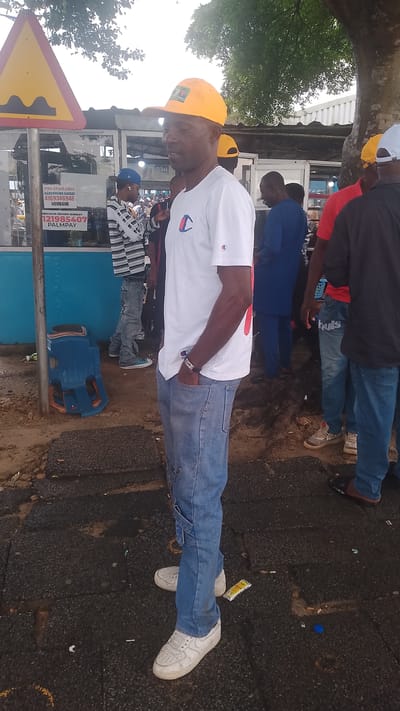 A man wearing a yellow cap and a white t-shirt with a red and blue logo stands with his right hand in his pocket. He is wearing blue jeans and white sneakers. He is in a public area, possibly a marketplace, with other people visible in the background. There is a speed bump sign on a pole to the left of the frame, and a blue plastic chair is overturned on the ground. The ground is paved with asphalt and dirt. The environment appears to be outdoors, with trees and a cloudy sky. The location is Asubi, Nigeria.