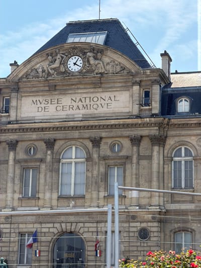 The image shows the facade of a large, ornate building in Sèvres, France. The building is made of stone and features classical architectural elements such as columns, pilasters, and carved reliefs. A prominent clock with Roman numerals is set into an arched recess above the main entrance. The text "MUSÉE NATIONAL DE CERAMIQUE" is inscribed on the building's facade.
The building appears to be a museum, likely dedicated to ceramics, given the inscription. The clock indicates it is approximately 10:50. The sky is visible in the upper left corner, suggesting it is daytime, and the lighting indicates it is a sunny day. The facade is adorned with sculptures and decorative elements, including figures and foliage. Small French flags are visible near some of the windows on the right side of the building. The overall impression is of a grand and historic public building.