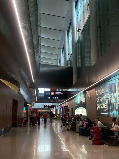 The image depicts the interior of an airport, likely in a city with a Spanish-speaking population based on the advertising signs. Passengers are seen both seated with their luggage and walking along a corridor. Airport signage, "Embarque Internacional" is visible. The architecture includes high ceilings, glass panels, and modern lighting fixtures. The floor appears to be polished tile. The scene suggests a typical airport environment with people waiting or moving through the terminal. There is no direct evidence to confirm the location in Canning, Argentina.