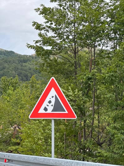 A triangular road sign with a red border and a white background is displayed. Inside the sign, a black silhouette depicts falling rocks. The sign is mounted on a metal pole and is positioned in front of lush green trees and a forested hillside. The sky above is overcast and grey. In the foreground, a section of a silver metal guardrail is visible. The image is set in a natural, mountainous environment, consistent with the outskirts of a city like Praga, Montenegro.