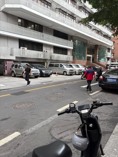 In the Liwan District of China, a street scene unfolds with a multi-story building featuring white latticework on its balconies and large glass windows on the ground floor. A row of parked vehicles, including silver SUVs and dark sedans, lines the street in front of the building. Several individuals are present: a young man in a black t-shirt and pants walks on the sidewalk, while an older man in a red and blue shirt and dark pants walks across the street. Two people are seen on a motorcycle in the background, and another individual is visible near the building entrance. A black electric scooter with a helmet hanging from its handlebars is prominently positioned in the foreground, facing towards the viewer. The street is paved and marked with yellow and white lines, with manhole covers visible on the asphalt. Trees with green foliage overhang the scene.