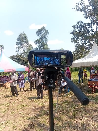 A black video camera mounted on a tripod stands in the foreground, angled to the right, with its screen displaying a blue image. A white cable descends from the camera setup. The scene depicts an outdoor gathering of approximately 15-20 individuals in the Butali Chegulo ward, Kenya.

In the mid-ground, people are distributed across a patchy grass and dirt field. On the left, a multi-colored tent (pink, light blue, white) provides shade, under which some individuals are seated. A man in a light-colored shirt stands near a large, light-brown cylindrical drum. Further right, a group of men in various shirts and trousers are standing, one holding a document. On the right side, a white tent is present, next to which a decorated wooden casket, adorned with red and white floral arrangements, is visible. Several other individuals, some seated, are near the white tent, including a person in a pink headscarf.

The background features numerous tall green trees, some with slender trunks, against a clear blue sky with scattered white clouds. The ground is uneven, showing areas of both sparse green grass and exposed brown soil. The overall environment is open and semi-rural.