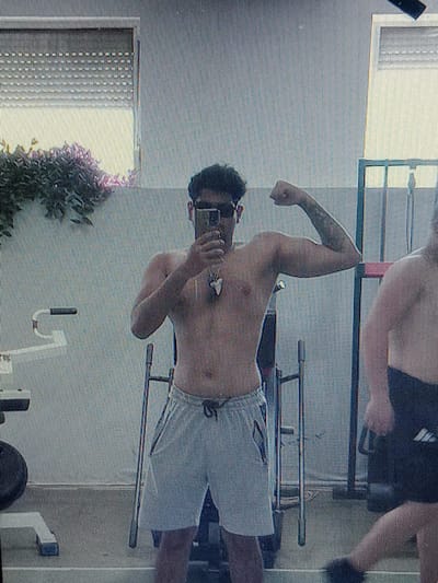 A full-body mirror selfie captures a shirtless man with dark hair and sunglasses posing in what appears to be a home gym or workout area. He is flexing his right bicep, holding a cell phone in his left hand to take the photo. He is wearing light-colored athletic shorts. Behind him, a window with blinds is visible, and to his right, another person is partially visible, also shirtless and wearing dark athletic shorts. The background suggests a residential setting, possibly a garage or a dedicated workout space. The image quality is grainy, indicative of a low-resolution camera or a screenshot. The location is identified as Gorno Aleksandrovo, Bulgaria.
