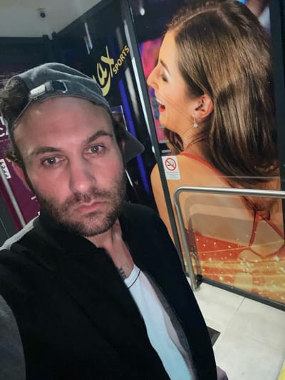 The image captures a selfie taken by a man indoors, likely in Newry, United Kingdom, with a large advertising display behind him. The man, in the foreground, looks directly at the camera with a neutral expression. He is wearing a dark hoodie and a grey baseball cap worn backward. A tattoo, partially visible on his inner forearm, appears to read "HER" or part of a longer word.
In the background, a vibrant advertising display features a smiling woman with long brown hair, wearing a red strappy top. She has a hoop earring in one ear and a dangling earring in the other. A metal railing runs horizontally across the display. A small white "NO SMOKING" sign with a red circle and cigarette pictogram is affixed to the display near the woman's shoulder. Above the man's head, the inverted text of a logo reads "3X SPORTS", suggesting a sports-related venue or advertisement. The floor is visible with light-colored tiles. The setting appears to be a commercial interior with artificial lighting.
