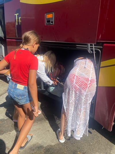 In Pol. Ind. La Palmosa, Spain, two individuals are interacting with the storage compartment of a maroon bus with yellow accents. On the left, a young person with blonde hair pulled into a ponytail wears a red t-shirt and denim shorts, holding a white object, possibly food, and a phone. To the right, another person, partially obscured by a white, sheer cover-up, wears a red bikini and is reaching into the bus's compartment. Several bags, including a patterned one, are visible inside the compartment. The ground is paved.