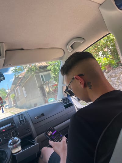 The image depicts a male individual seated in the passenger side of a vehicle, viewed from behind and slightly to the right. He has a faded haircut and a black rose tattoo on the right side of his neck. Dark sunglasses are perched on his ear. He is wearing a black t-shirt and holds a smartphone with both hands, its screen displaying a dark-themed application, possibly a messaging app, with a visible timestamp of "13:36". A partial tattoo is also visible on his right thigh.

The car's interior includes a light-colored fabric headliner, a gray dashboard with a Sony car radio in the center console, and an open storage compartment below it revealing USB cables. An iced coffee drink in a clear plastic cup with a straw rests in a cup holder. A decorative item hangs from the rearview mirror.

Beyond the windshield, a residential street scene unfolds under a clear blue sky. A light grey house with a red-tiled roof and white windows dominates the view, surrounded by lush green trees and foliage. Further down the street, another building and a small grey car are visible. The scene is located in Касапите, Bulgaria.
