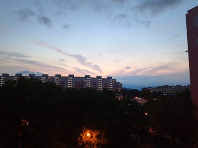 An exterior, eye-level, medium shot captures a dusky sky with wisps of pink and grey clouds above a long, multi-story apartment building in the city of Most, Czechia. The apartment building, which spans the middle ground, is dimly lit with windows illuminated by interior lights. In the foreground, dark silhouettes of trees obscure the lower half of the frame, with a single streetlamp casting an orange glow through the foliage. To the right, the edge of another building, a tall, reddish-brown structure with circular cutouts, is visible. The scene suggests early evening or twilight in a residential area.