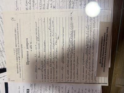 This is a close-up, eye-level, indoor shot of a document with handwritten Russian text. The document appears to be an official record, possibly an interrogation protocol or personal file, based on the headings and numbered questions. The document is cream-colored and shows signs of age, with some creasing and discoloration. A bright white circular light source is reflected in the upper right quadrant of the image, obscuring some of the text. A portion of another document, also with Russian text, is visible at the bottom right, featuring a stamp that reads "Дело политзаключенного АНДРЕЕВА НИКОЛАЯ ИОСИФОВИЧА расстрелянного в Акхунском лагере НКВД". The main document has a title "ПРОТОКОЛ ДОПРОСА" (Interrogation Protocol) at the top. Below this, it is identified as originating from "НАРОДНЫЙ КОМИССАРИАТ ВНУТРЕННИХ ДЕЛ" (People's Commissariat of Internal Affairs) and a specific department "Отдел Охраны ИТЛ МЗ УНБД по Новосибирской области" (Department of Protection of ITL MZ UNBD for Novosibirsk Oblast). The date "1938 г. февраль" (February 1938) is present. Several questions are numbered, and some have handwritten answers. For example, question 1 asks for "Фамилия" (Surname), and the answer is written as "Андреев". Question 2 asks for "Имя и отчество" (First name and patronymic), with the answer "Николая". Question 3 is "Дата рождения" (Date of birth) with "1896 г." (1896). Question 4 asks for "Место рождения" (Place of birth) and has the answer "Баш. АССР." (Bashkir ASSR). Question 5 is "Местожительство" (Residence) with the answer "г. Аханькское." (Ahan'kskoe). Question 6 asks for "Нац. и граж. (подданство)" (Nationality and citizenship (allegiance)) with the answer "на агари" (on agar - this part is unclear). Question 7 is "Паспорт" (Passport) with a handwritten "пр" (likely an abbreviation). Question 8 asks for "Род занятий" (Occupation) with the answer "общир род. (ком)" (general occupation - unclear). Question 9 is "Социальное происхождение" (Social origin) with the answer "Сибиряк" (Siberian). Question 10 asks for "Социальное положение (род занятий и имущественное положение)" (Social status (occupation and property status)), and question 11 asks for "Состав семьи" (Family composition) with the answer "у родителей, на родителей и их имущественное положение" (for parents, for parents and their property status). Some additional handwritten notes are present in the margin and between lines, including "гр. 34 г. гражданин" (citizen of 34 years) and "родственники, из им" (relatives, from them). The location of Ust'-Kabyrza, Russia is not explicitly mentioned in the visible text of this document. However, the document relates to