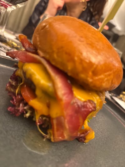 A close-up shot displays a gourmet burger. The burger features a glazed bun, melted cheese, several slices of crispy bacon, and lettuce. It seems to be placed on a dark grey plate and it is skewered together. The photo is taken at a slight angle, giving an upper perspective on the burger. A person can be seen holding a beverage in the background.