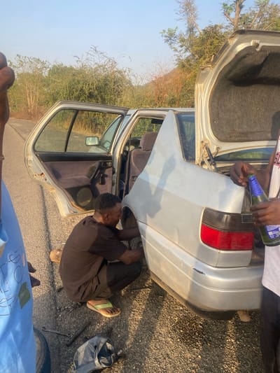 The image depicts an outdoor scene in the city of Gu, Nigeria, situated on a paved asphalt road adjacent to an area with dense, predominantly green and dry vegetation, backed by hilly terrain. A stationary silver four-door sedan, identified as a Volkswagen Jetta type vehicle due to its taillight and body design, is present. Its rear passenger-side door is ajar, exposing a dark fabric interior, and its trunk lid is open. A dark-skinned male subject, dressed in a black t-shirt, black trousers, and light green flip-flops, is crouched at the rear passenger-side wheel well, actively engaged in mechanical work, likely a tire change or repair. Various hand tools, including wrenches, are scattered on the asphalt directly in front of him, next to a black bag. To the far left of the frame, the arm and shoulder of another dark-skinned individual wearing a light blue garment are partially visible. To the right, the right arm and torso of a third dark-skinned person in a white t-shirt are visible, holding an open green glass bottle with a light blue cap. The scene is illuminated by clear daylight, creating distinct shadows. The vehicle exhibits minor wear, including a scratch on the rear bumper.